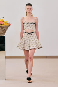 Model walking a runway in a cream butterfly-print two-piece set: cropped top with black trim and a pleated mini skirt, gold earrings, and black sandals.