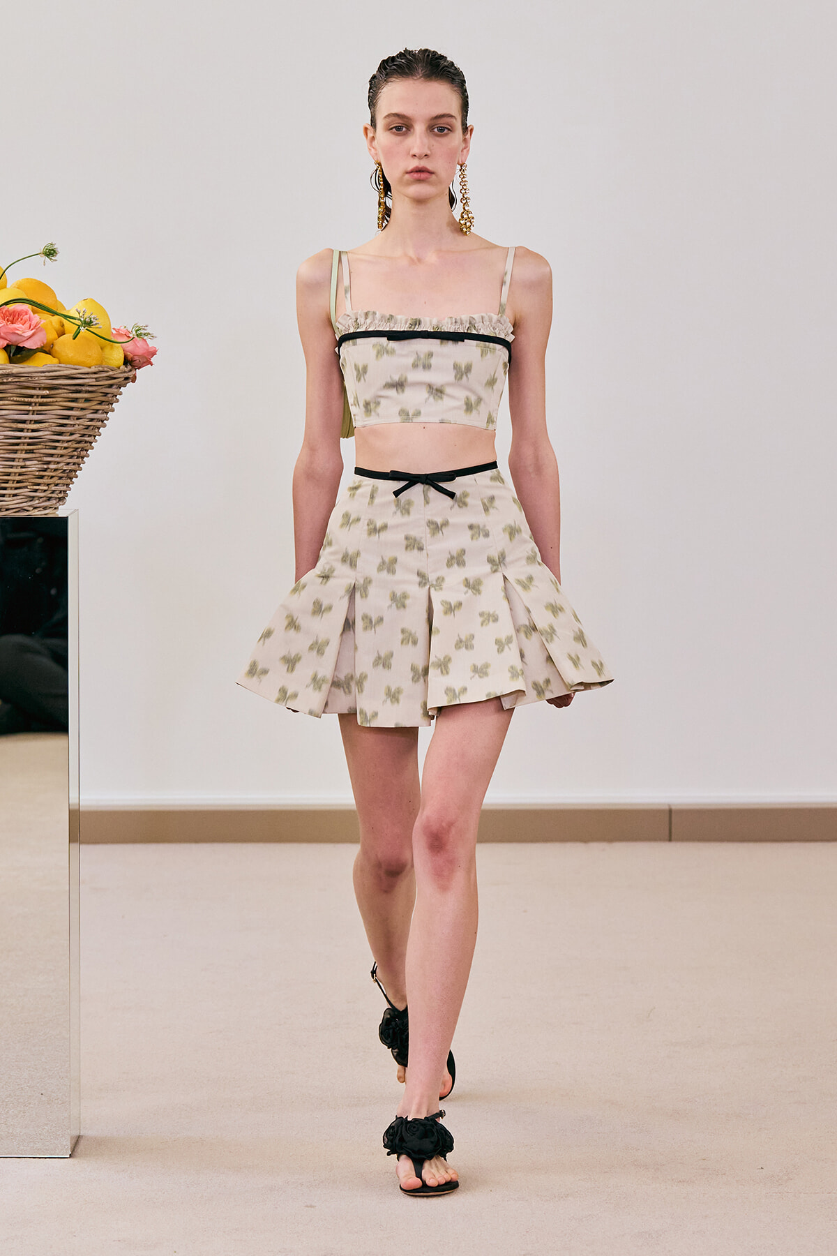 Model walking a runway in a cream butterfly-print two-piece set: cropped top with black trim and a pleated mini skirt, gold earrings, and black sandals.