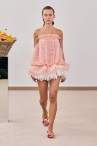 Model walking a pink strapless ruffled mini dress with white feather trim on a fashion runway, wearing pink flower sandals.