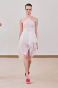 Model walking a runway in a white lace halter dress with scalloped edges and an asymmetrical high-low hem, paired with pink flower-embellished heels.
