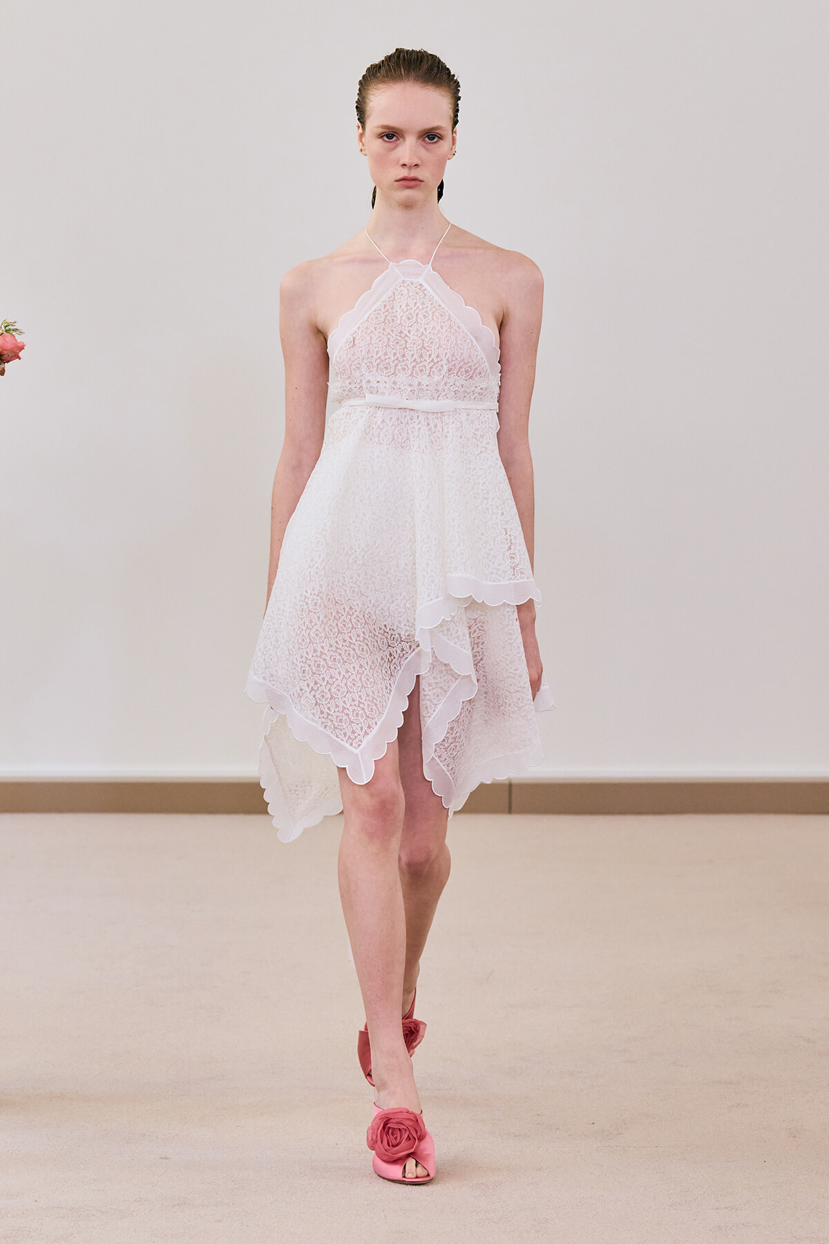 Model walking a runway in a white lace halter dress with scalloped edges and an asymmetrical high-low hem, paired with pink flower-embellished heels.