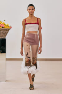 Model on a runway wearing a beige cropped top with red ruffle and a sheer floral-print skirt with a white feathered hem and floral-embellished heels.