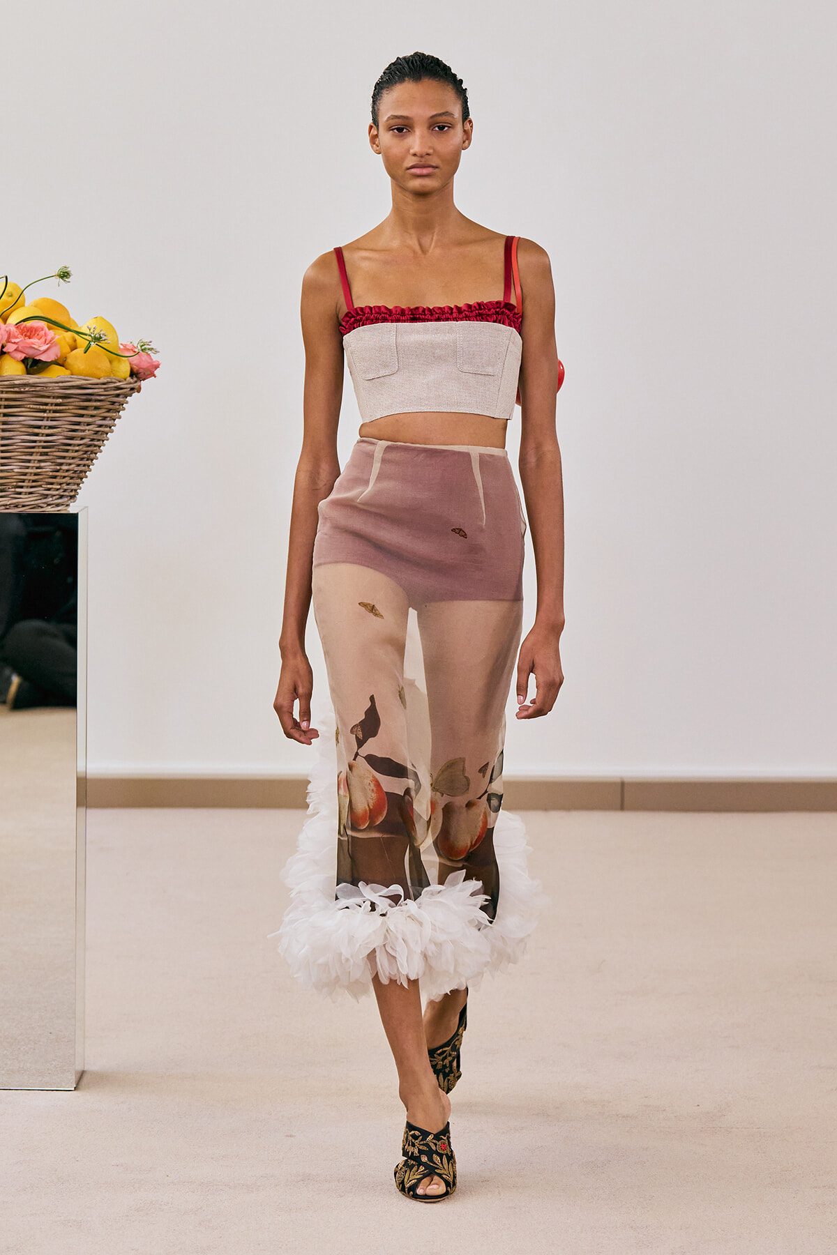 Model on a runway wearing a beige cropped top with red ruffle and a sheer floral-print skirt with a white feathered hem and floral-embellished heels.