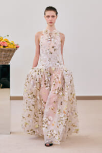 Model wearing a pale pink floral lace gown with a sheer, embroidered skirt and bow at the neckline in a studio setting.