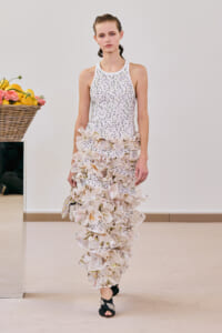 Model walking in a white, sleeveless dress with dotted pattern and tiered ruffle layers, holding a small clutch, indoors beside a basket of lemons and flowers.