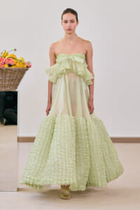 Model wearing a pale green strapless gown with multiple ruffled layers and a voluminous, textured skirt photographed indoors.