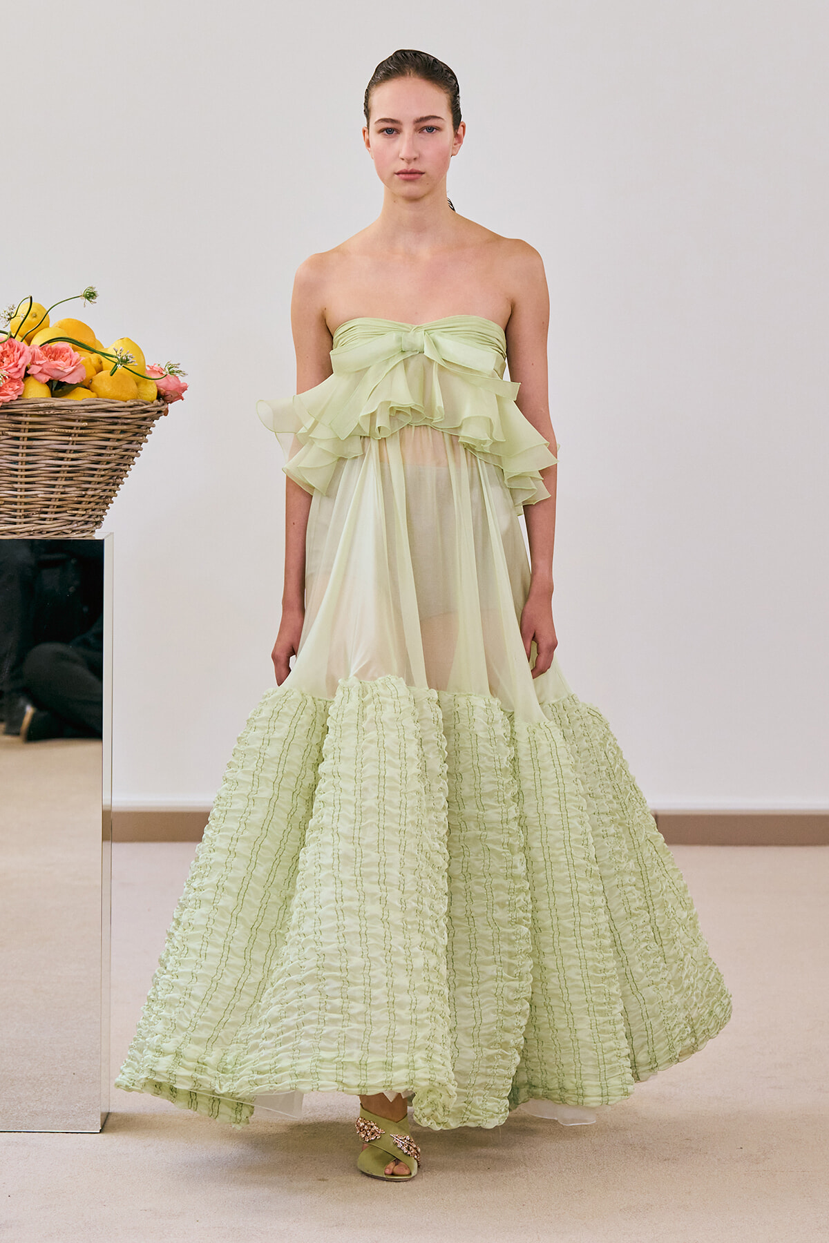 Model wearing a pale green strapless gown with multiple ruffled layers and a voluminous, textured skirt photographed indoors.