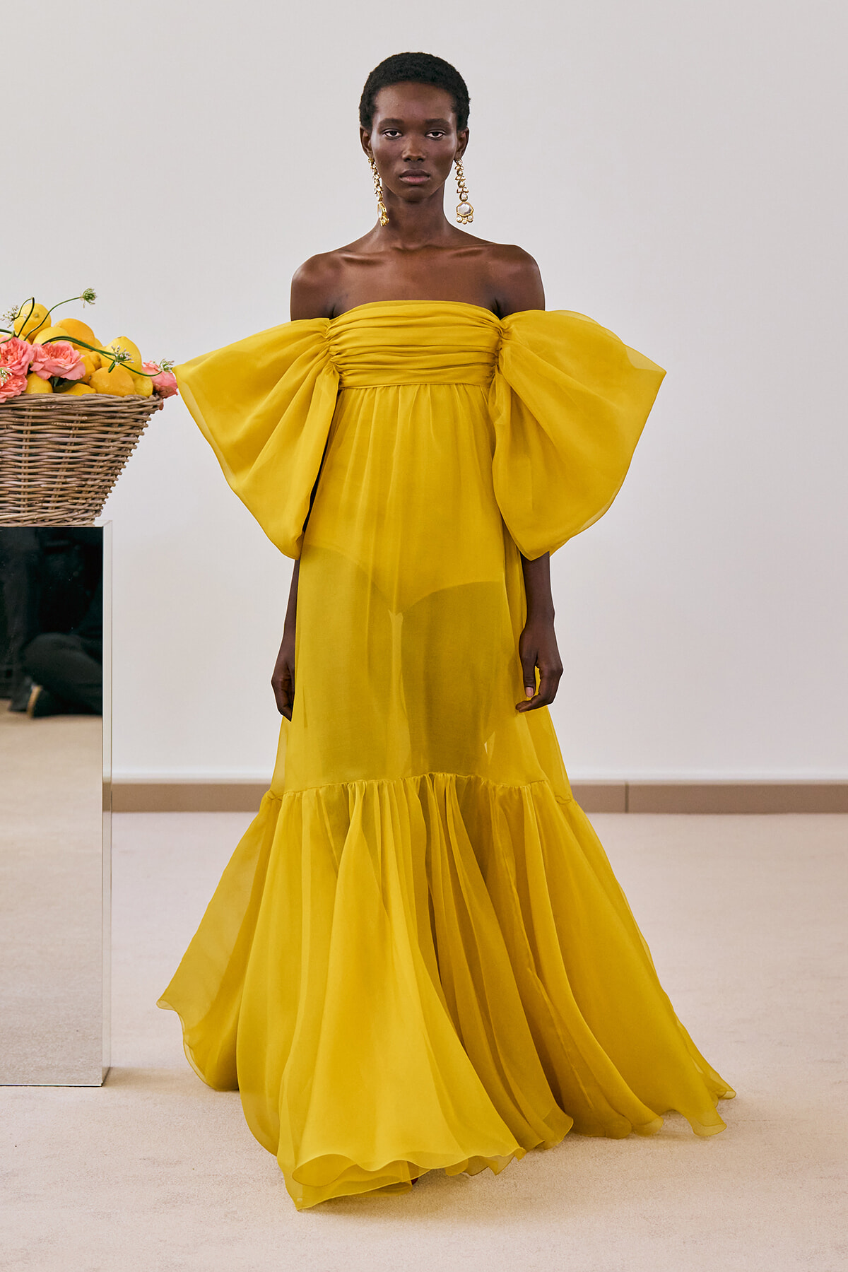 Model wearing a bright yellow off-the-shoulder gown with oversized puff sleeves, standing in a neutral room.