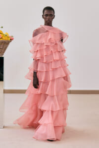 Model wearing a floor-length pink, multi-layered ruffle gown in a neutral room.