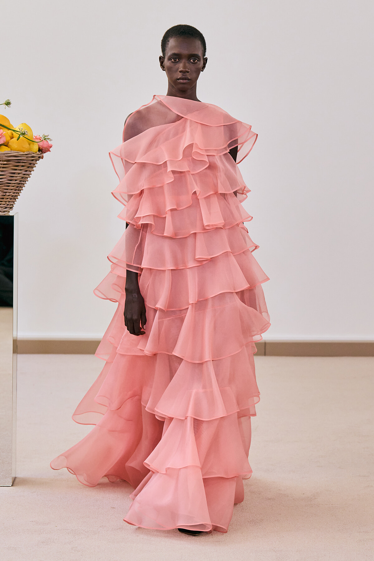 Model wearing a floor-length pink, multi-layered ruffle gown in a neutral room.