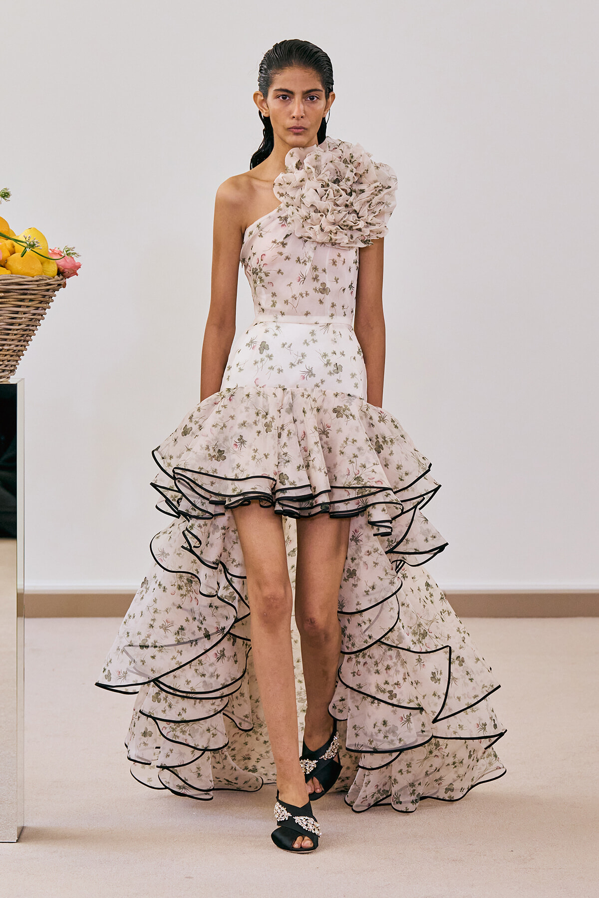 Model in a pale pink floral one-shoulder gown with a large shoulder flower and a tiered, black-trimmed ruffled skirt.
