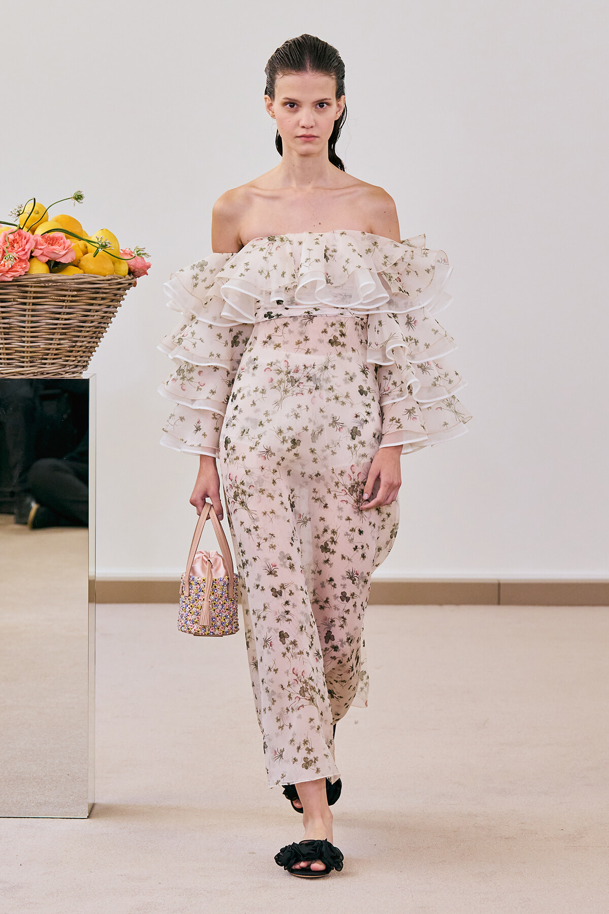 Runway model in a pale pink floral off-the-shoulder outfit with tiered ruffles, carrying a small pink beaded handbag beside a basket of lemons and flowers in a studio setting.