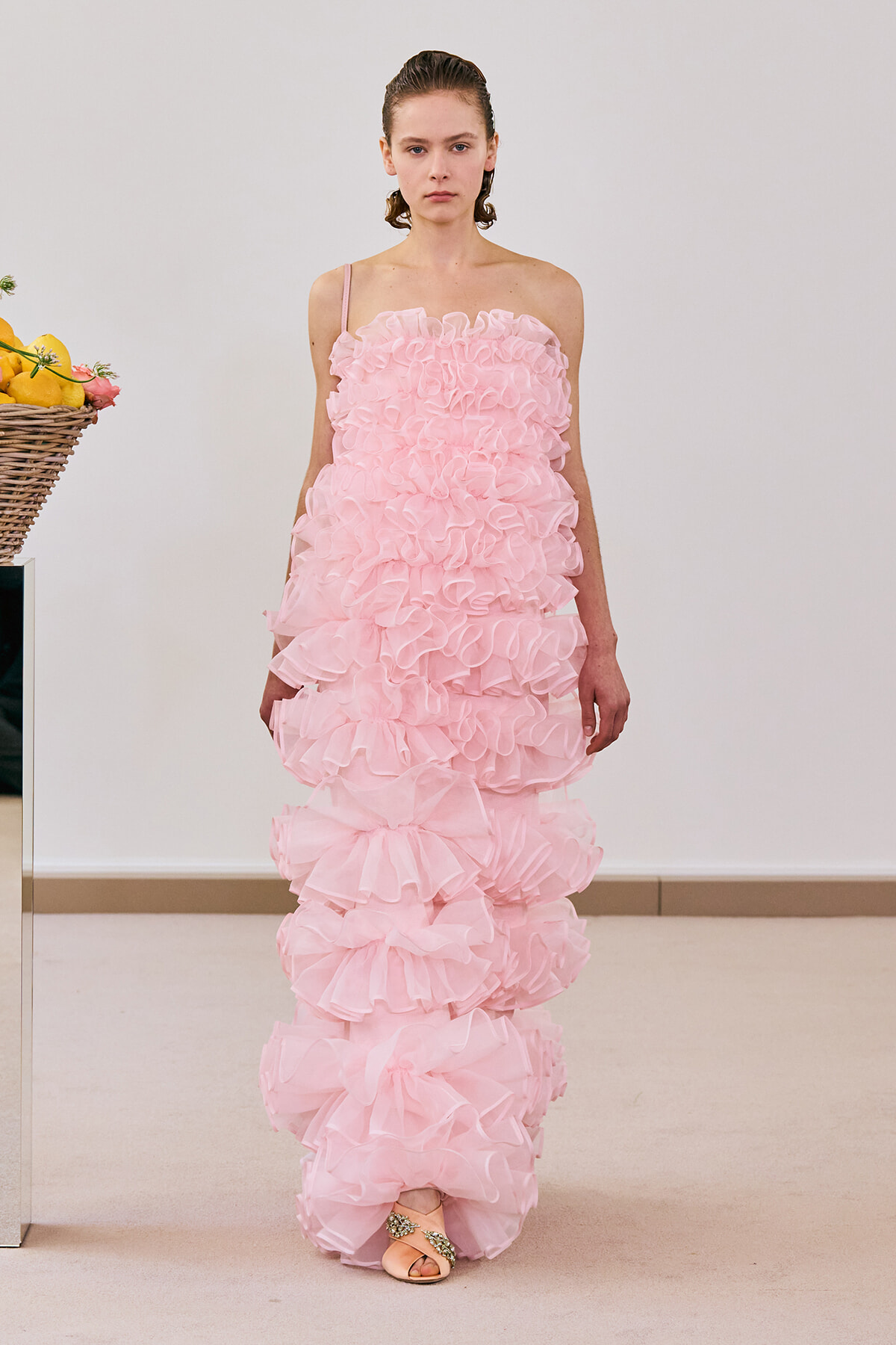 Model wearing a pale pink strap-detailed gown with cascading layered ruffles reaching the floor, in a studio setting.