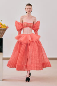 Model in a coral pink off‑shoulder top with large puff sleeves and a voluminous tiered skirt, walking on a runway.