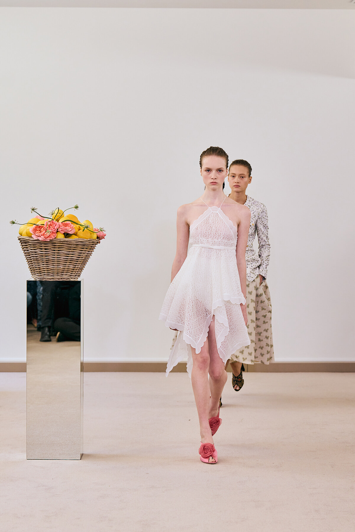 Female model in a white lace halter dress walks a runway, with a wicker basket of lemons and pink flowers atop a mirrored pedestal nearby.