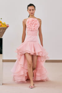 Model wearing a pale pink halter gown with a large ruffled flower bodice and a layered high-low skirt in a studio setting.