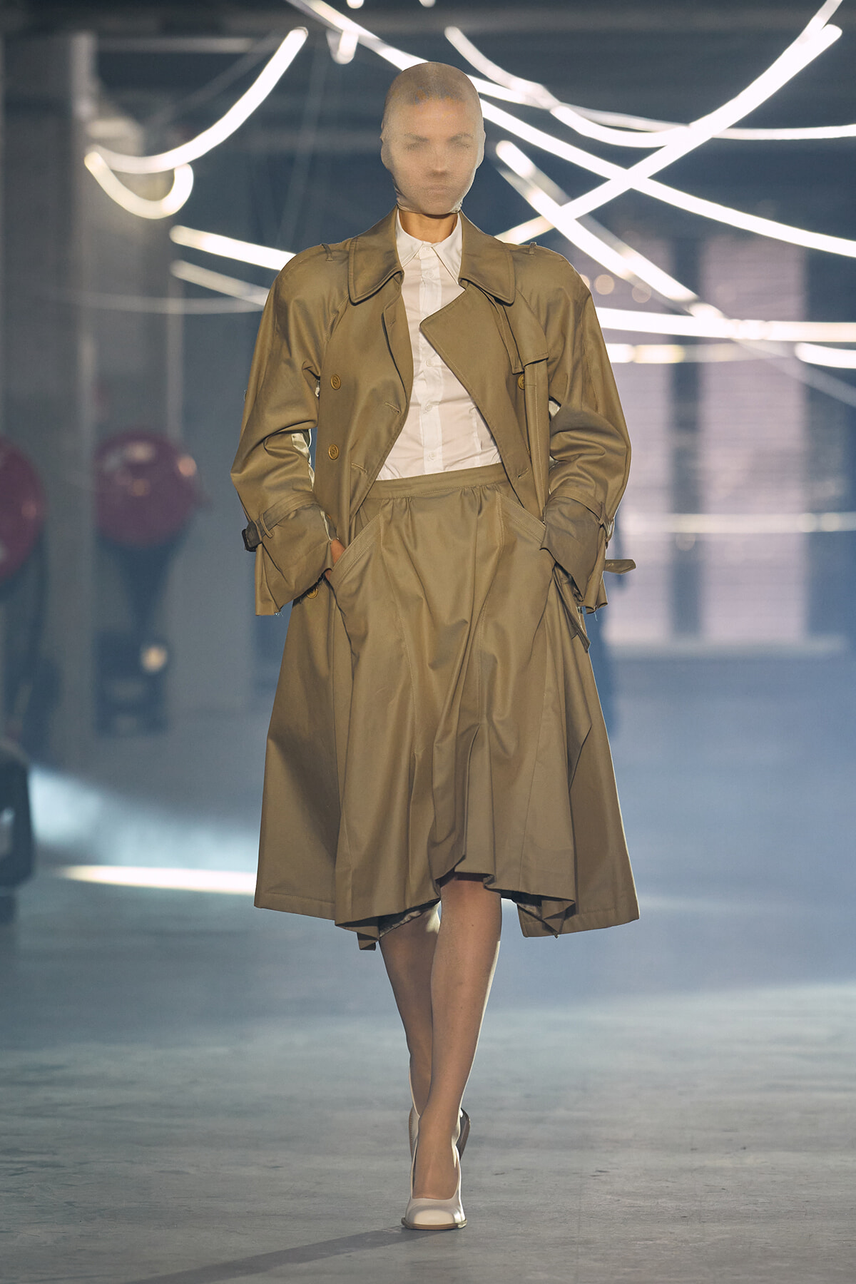 Model walks the runway in a tan trench coat over a white shirt and midi skirt, hands in pockets, white heels.