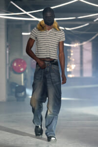Masked male model walking a fashion runway in a striped polo with a fur-curred collar and baggy jeans in an industrial setting