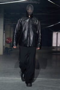 Model walking on a runway in an all-black outfit: oversized black leather zip jacket with high collar, wide black pants, black boots, in an industrial space.
