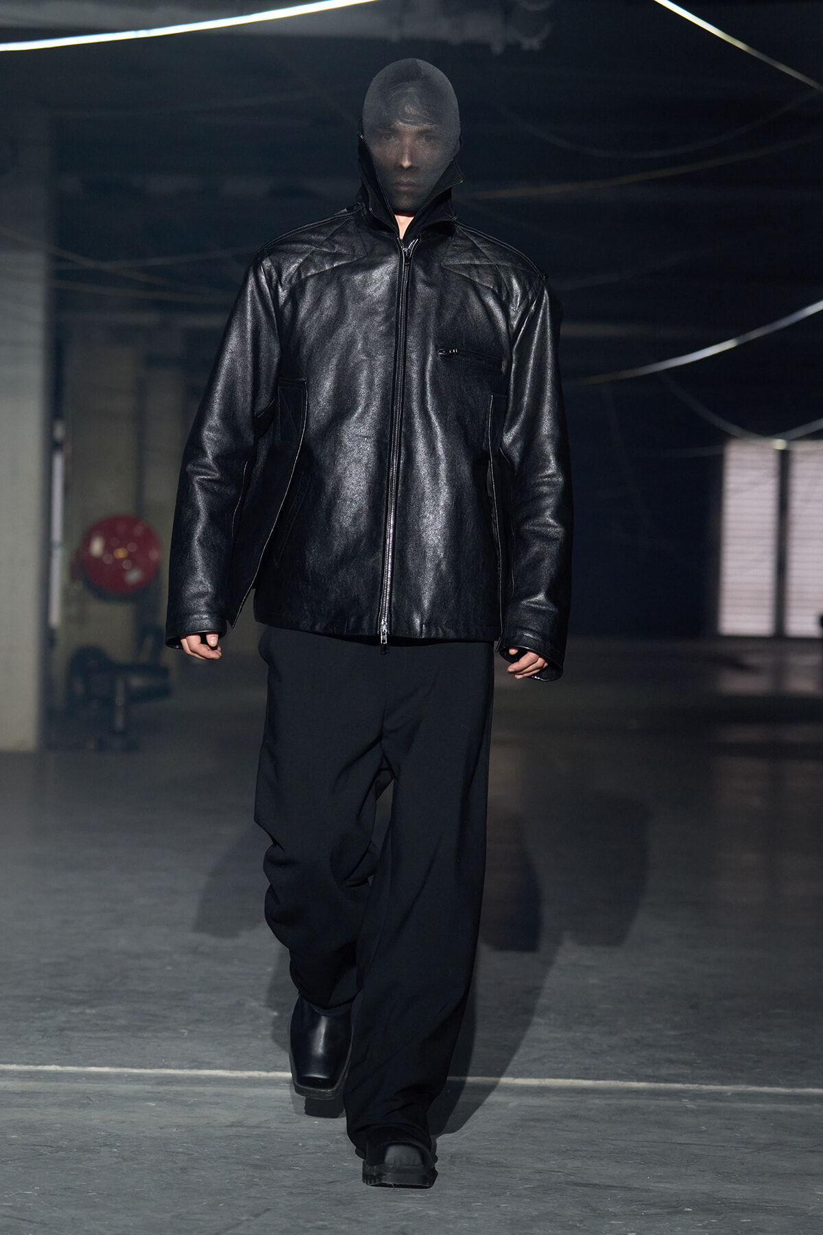 Model walking on a runway in an all-black outfit: oversized black leather zip jacket with high collar, wide black pants, black boots, in an industrial space.