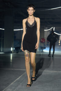 Model walking a runway in a short black lace-trim slip dress with halter neck, black heeled sandals, and visible tattoo on her left thigh.