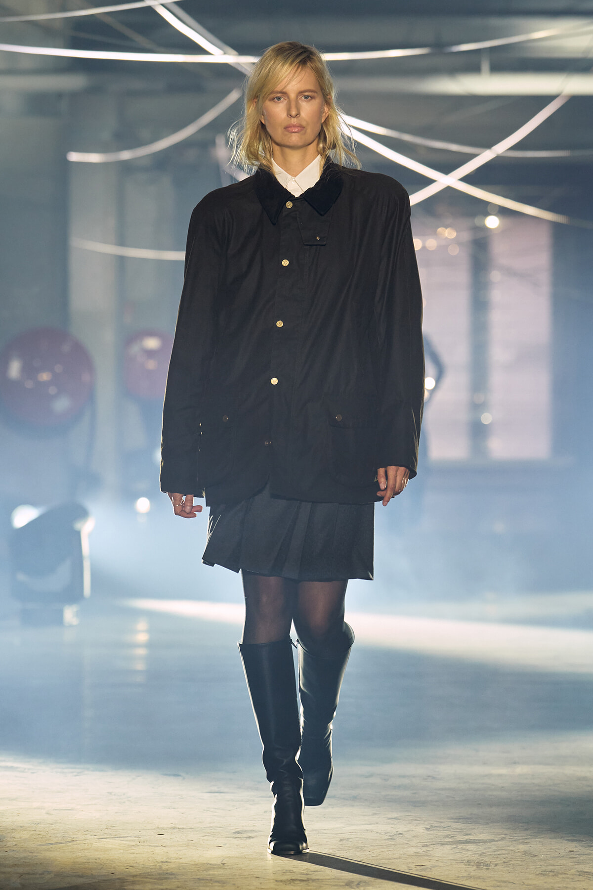 Female model walking a runway in an oversized black coat over a white shirt, paired with a short pleated skirt and knee-high boots under dramatic lighting.