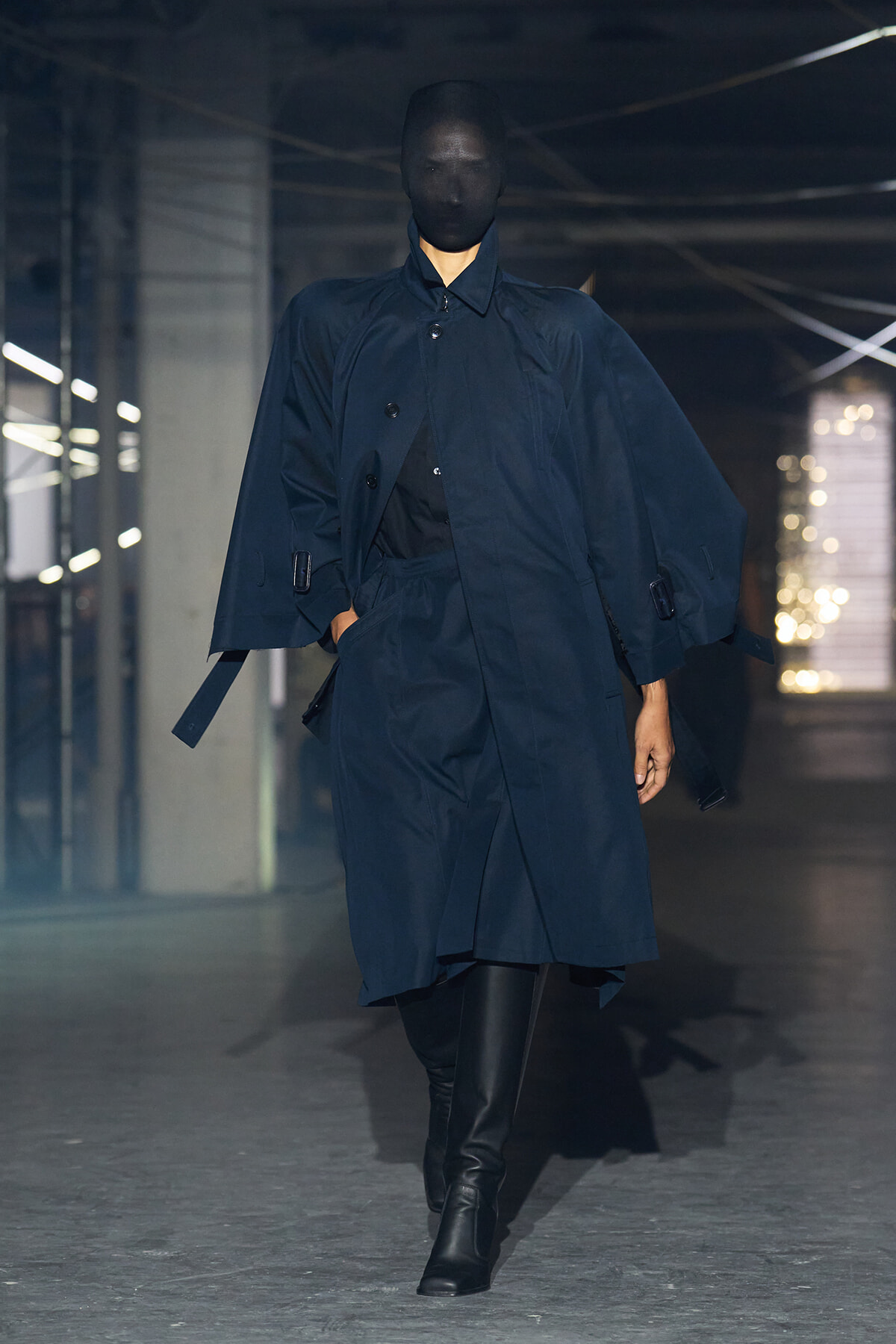 Model walking a fashion runway in a navy trench coat with a belt and black knee-high boots; face obscured by a dark mask in a dim, industrial setting.