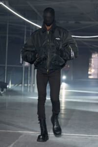 Model in a black oversized bomber jacket, masked face, black jeans and combat boots walking a dim fashion runway.