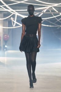 Model walks a fashion show runway in an all-black outfit: polo top, pleated mini skirt, tights, and black heels under bright neon lights overhead.