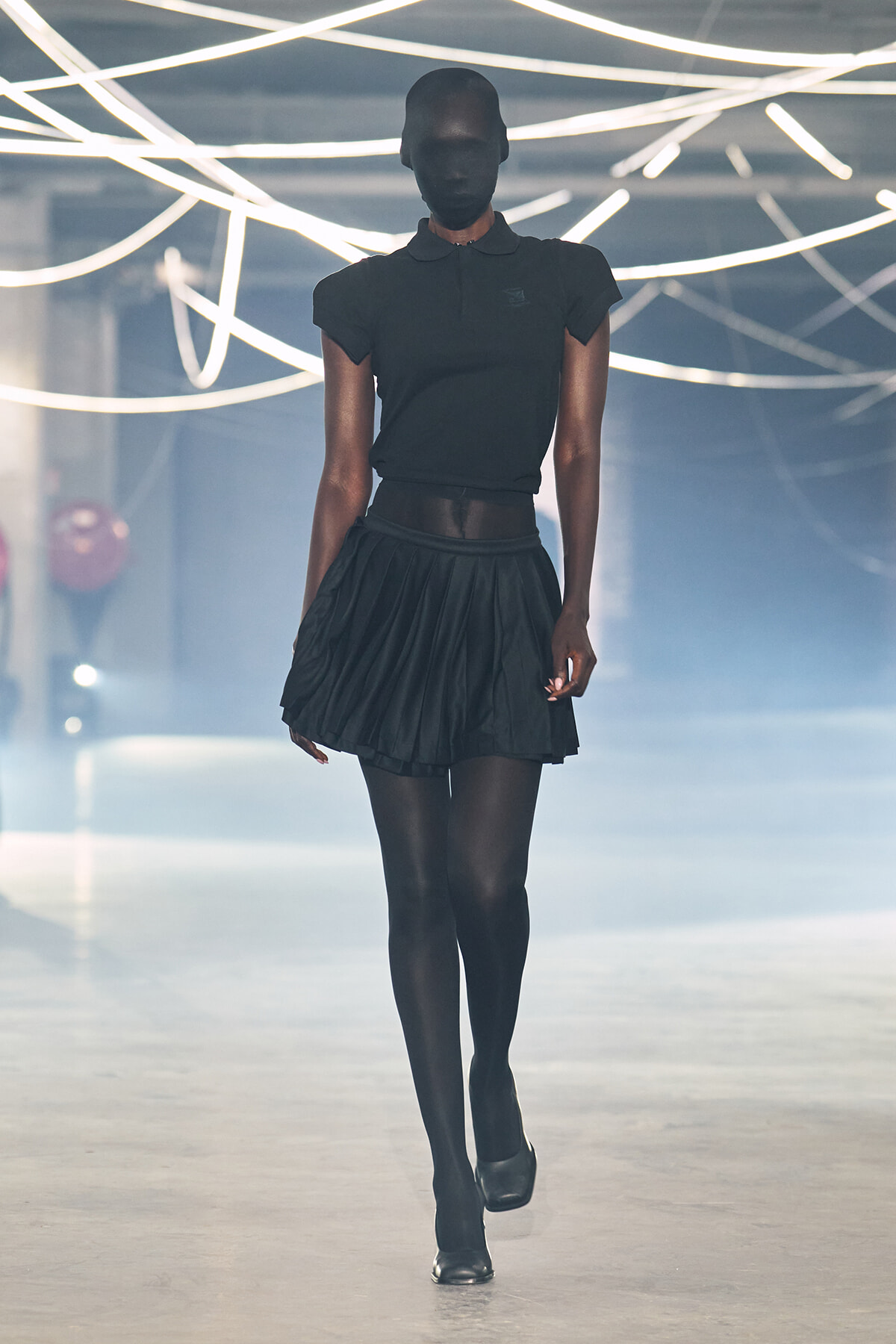 Model walks a fashion show runway in an all-black outfit: polo top, pleated mini skirt, tights, and black heels under bright neon lights overhead.