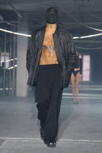 Male model walks a fashion runway in a black balaclava, open leather jacket, and wide black trousers; chest tattoos visible.