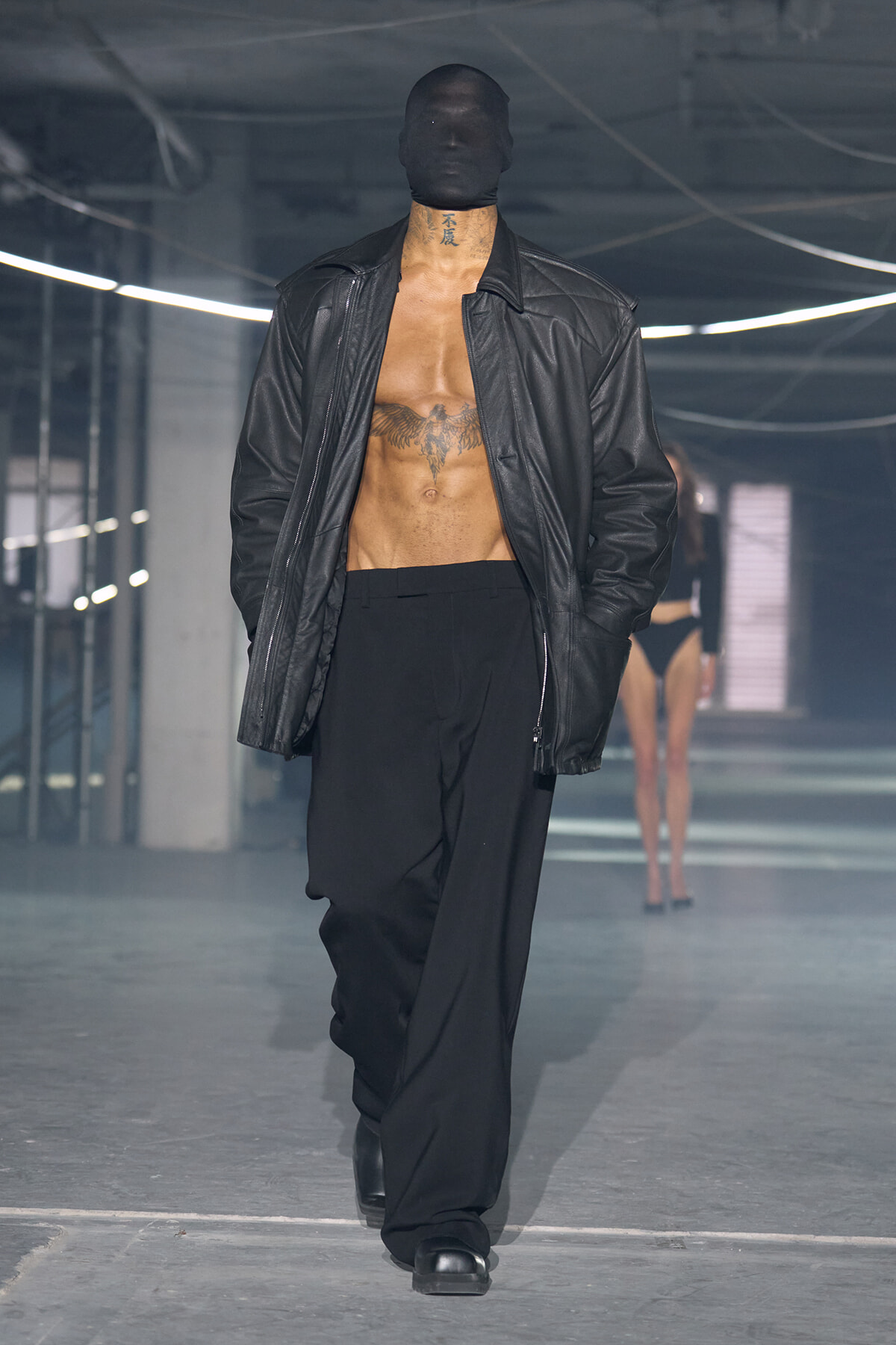Male model walks a fashion runway in a black balaclava, open leather jacket, and wide black trousers; chest tattoos visible.