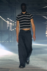 Model on a runway wearing a black balaclava, striped cropped polo, and dark cargo pants in a dim studio setting.