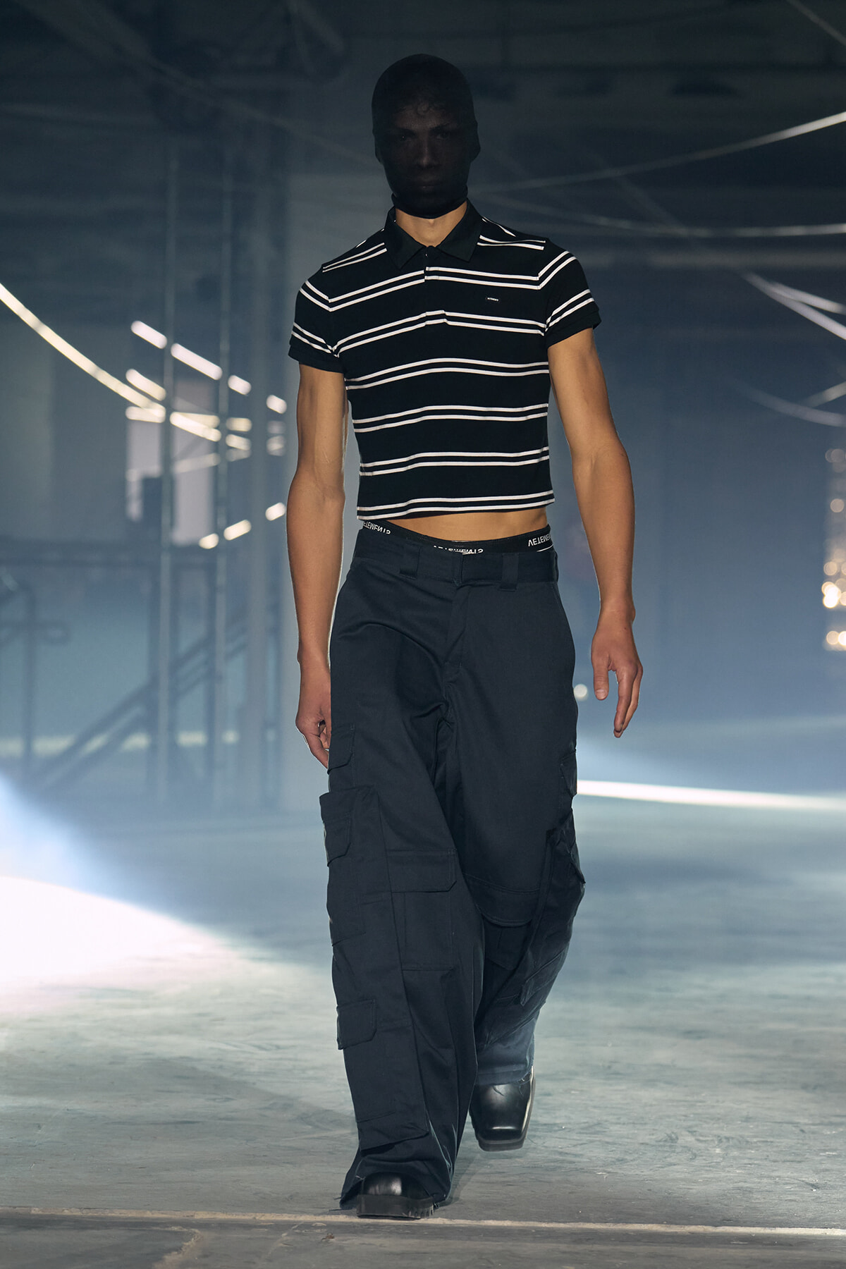 Model on a runway wearing a black balaclava, striped cropped polo, and dark cargo pants in a dim studio setting.