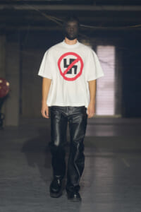 Male model walking on a runway in a white T-shirt featuring a red crossed-out swastika symbol.
