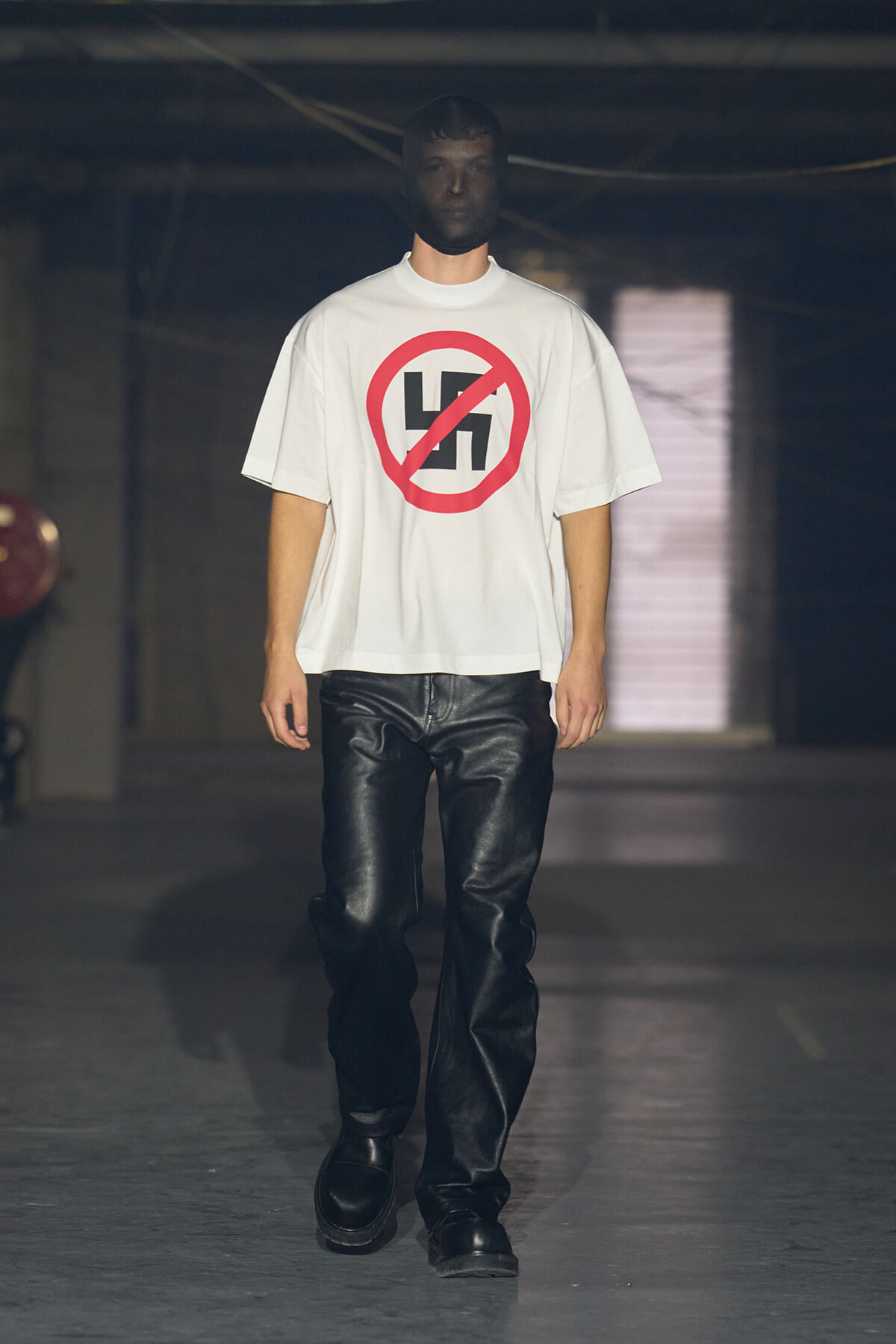 Male model walking on a runway in a white T-shirt featuring a red crossed-out swastika symbol.