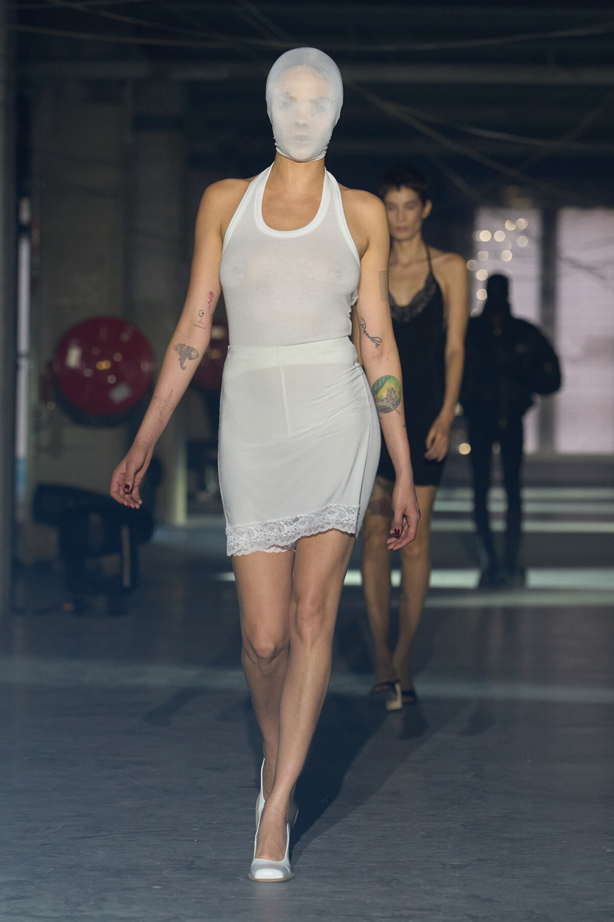 Model walking a runway in a white sleeveless dress with a sheer mask over her head; another model follows in black behind her.