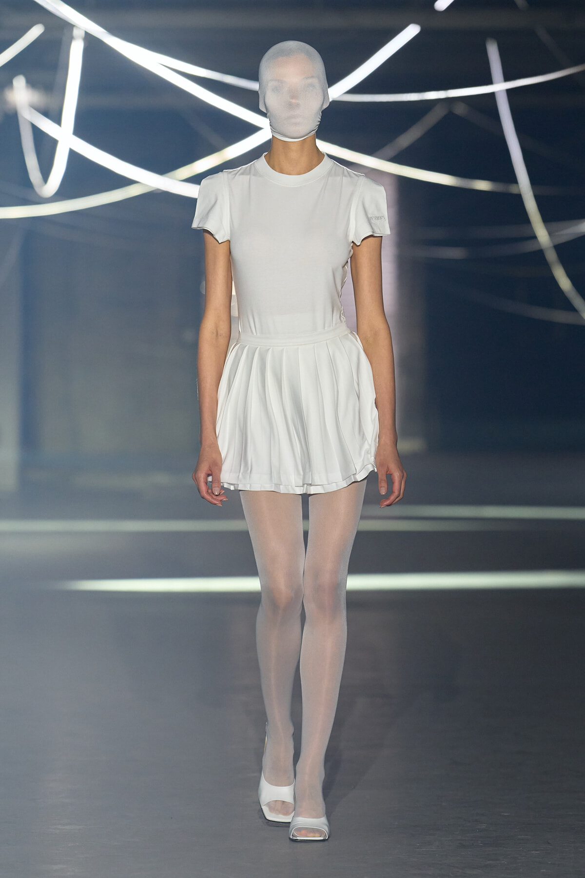 Model on a runway wearing an all-white outfit: fitted T-shirt, pleated mini skirt, sheer tights, white heels, and a white head covering.