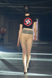 Model walking a runway in a black cropped T‑shirt with a red crossed‑out swastika, nude tights, and black heels; face covered by a black mask.