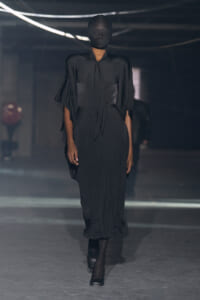 Model walks a dim runway in an all-black satin dress with draped cape sleeves and a head covering.