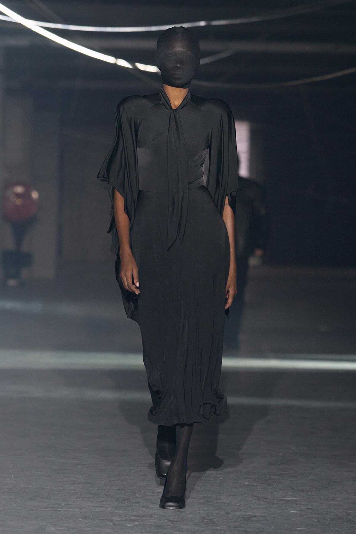 Model walks a dim runway in an all-black satin dress with draped cape sleeves and a head covering.