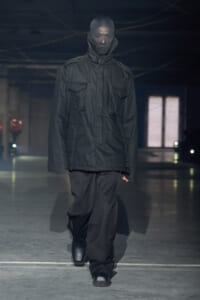 Model wearing a loose black utility jacket with large front pockets and a high collar, walking a dim runway in black pants and boots.
