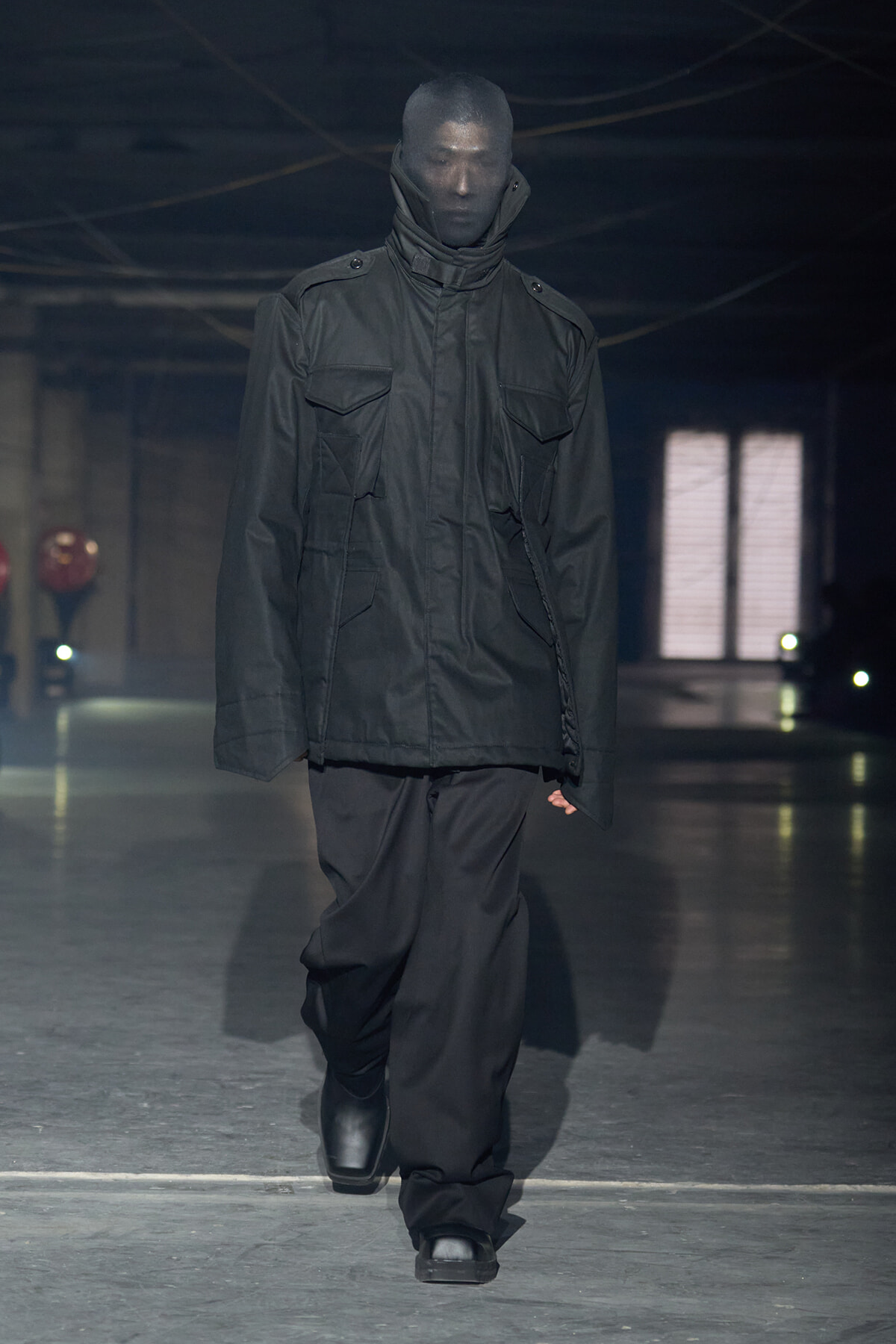 Model wearing a loose black utility jacket with large front pockets and a high collar, walking a dim runway in black pants and boots.