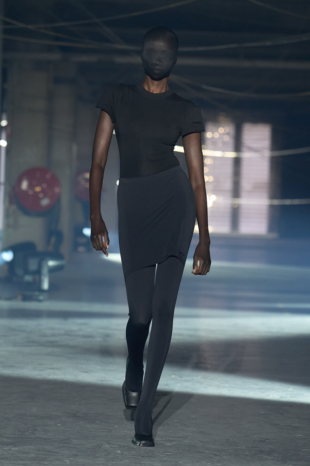 Model walks down a dim runway in an all-black outfit, with her face fully covered by black fabric under blue lighting.