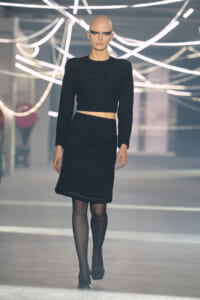 Model walking the runway in a black textured cropped jacket and matching knee-length skirt, bold makeup, and neon light installation in the background.