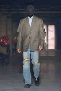 Runway model in an oversized gray blazer over a light blue shirt, distressed jeans, and black boots walking down the runway.