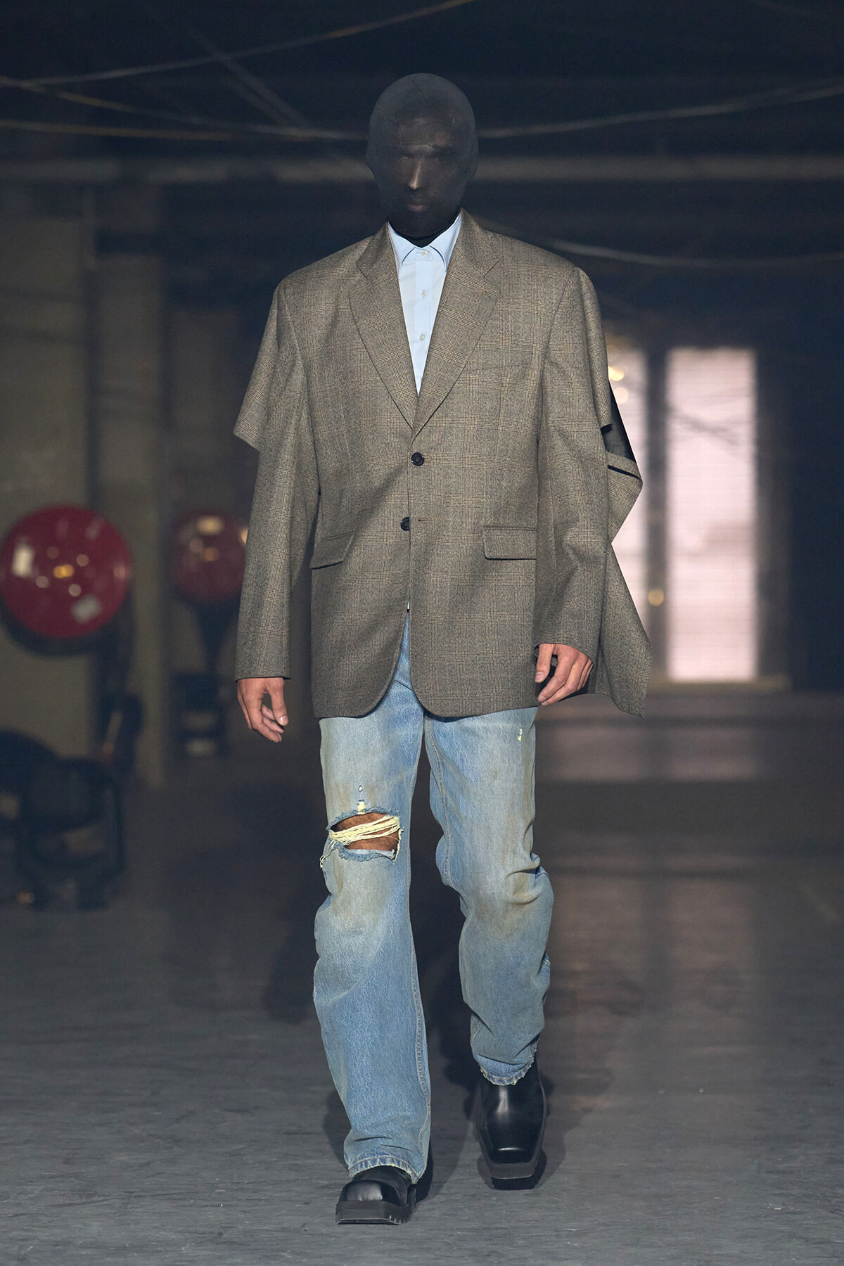 Runway model in an oversized gray blazer over a light blue shirt, distressed jeans, and black boots walking down the runway.