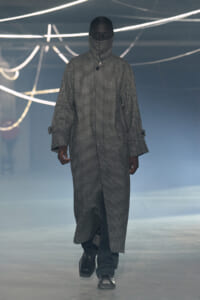 Model walking the runway in a long checkered coat with high collar and face covering, neon light backdrop creates a moody fashion scene