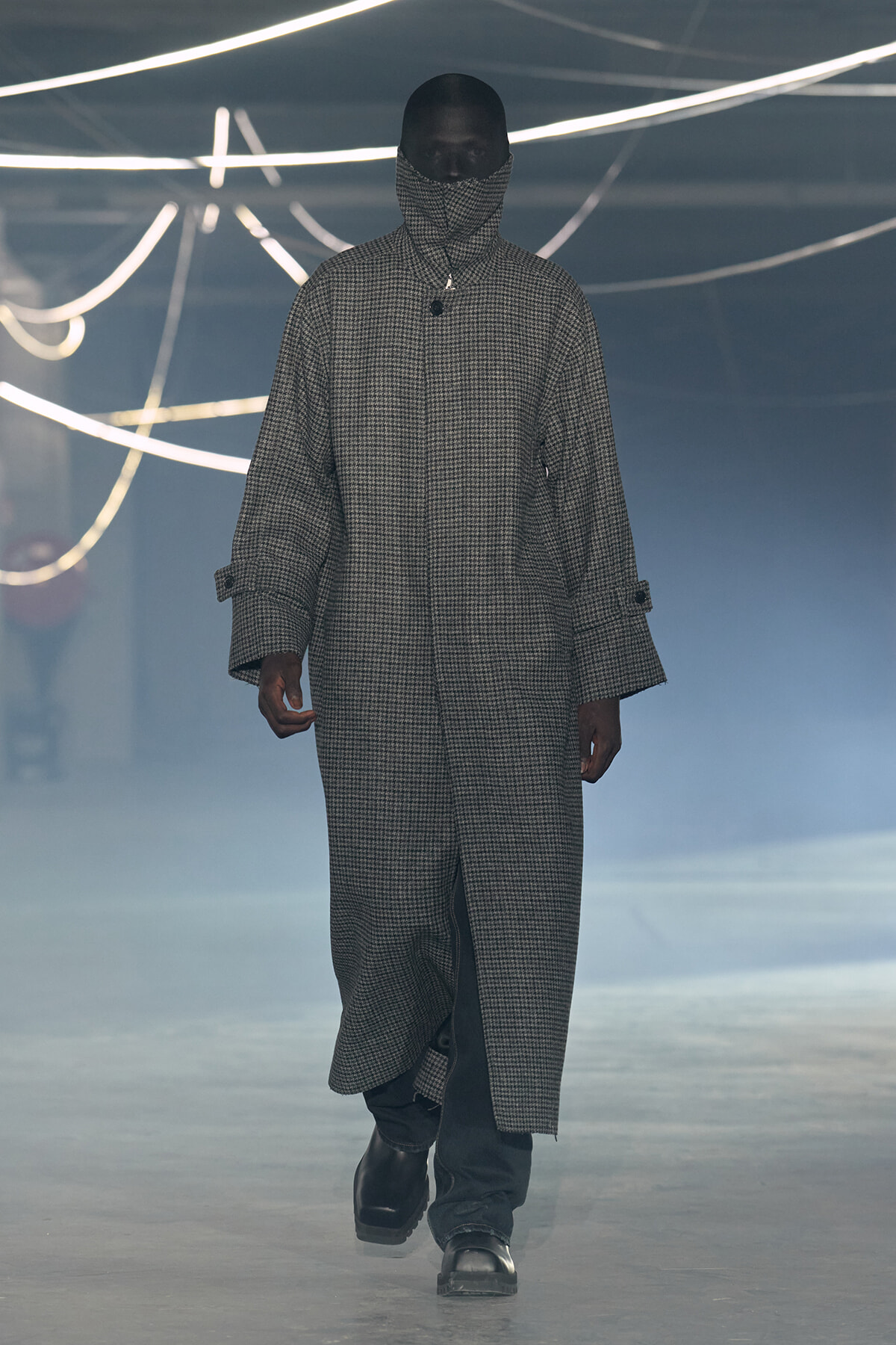 Model walking the runway in a long checkered coat with high collar and face covering, neon light backdrop creates a moody fashion scene