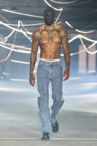 Tall shirtless male model with extensive chest and arm tattoos walking a fashion runway in blue jeans and a white Vetements waistband; neon light backdrop.